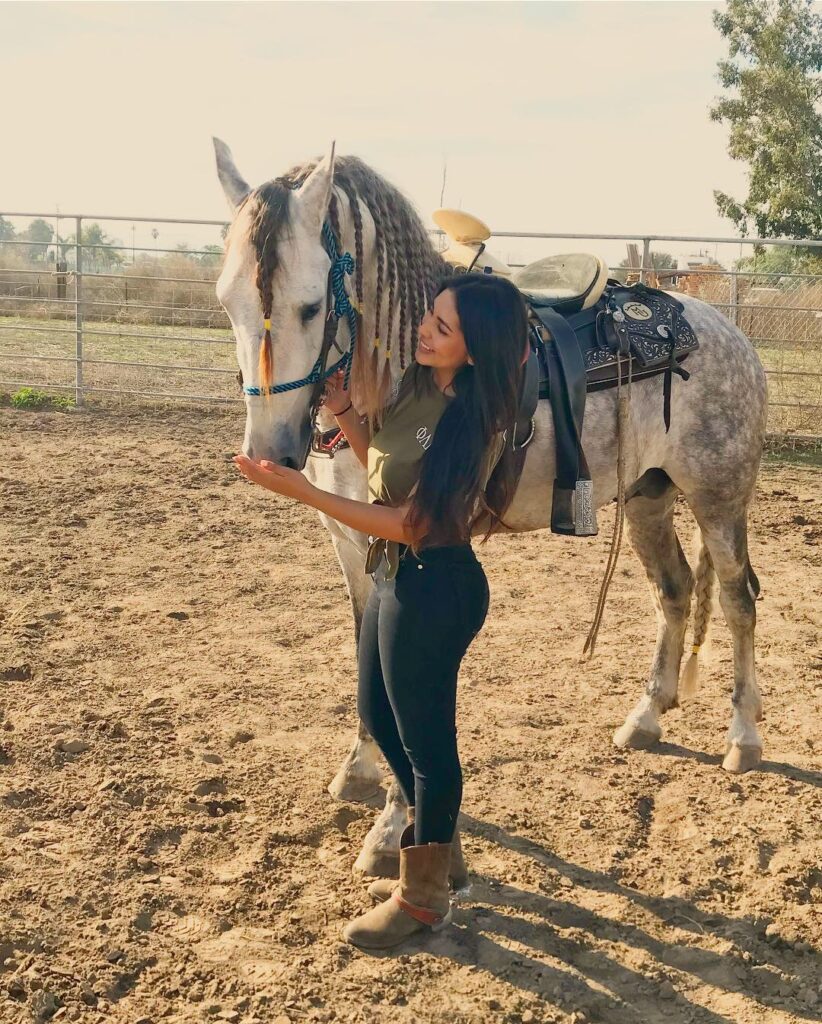 Women wearing cowgirl boots is looking at her horse that has a braided mane with a black saddle.